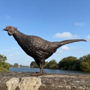 Bronze pheasant sculpture displayed beside a tranquil lake in warm morning sunshine.