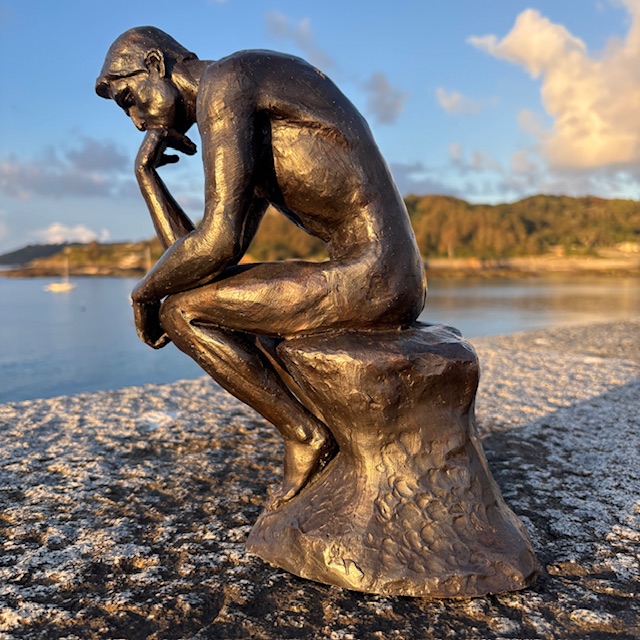 Bronze Thinker sculpture photographed at sunrise overlooking calm coastal waters, highlighting the figure's contemplative pose and rich hand-finished patina.