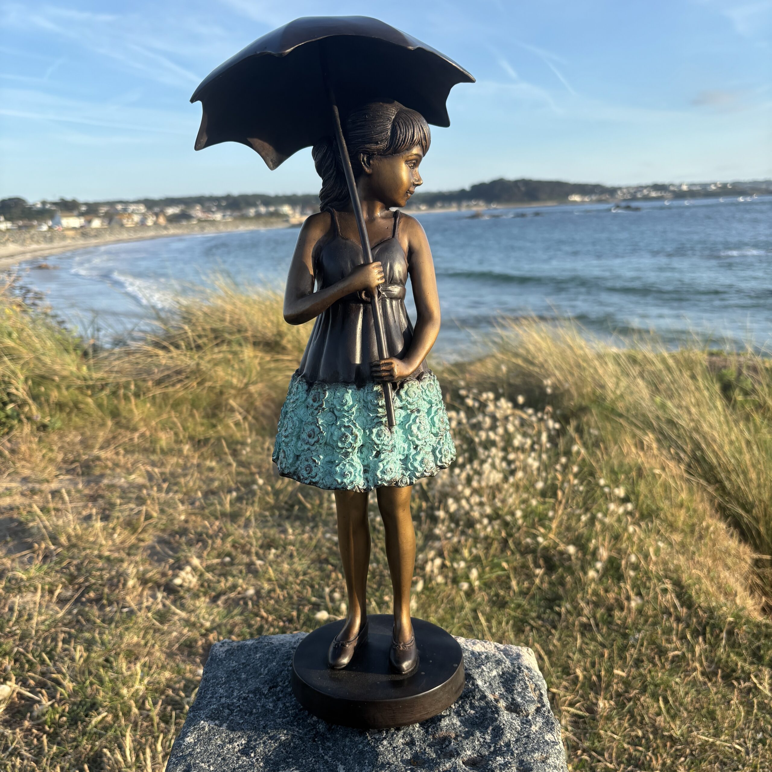 Bronze April Showers Sculpture Girl Holding Umbrella FIGI 72 full-length front view overlooking the sea and coastal landscape.