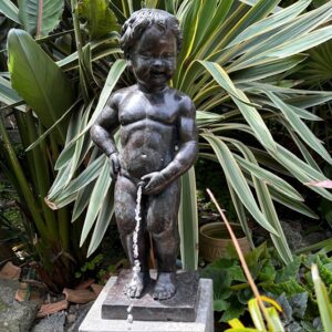 Bronze Manneken Pis fountain sculpture with flowing water, displayed on a stone pedestal among lush tropical garden foliage