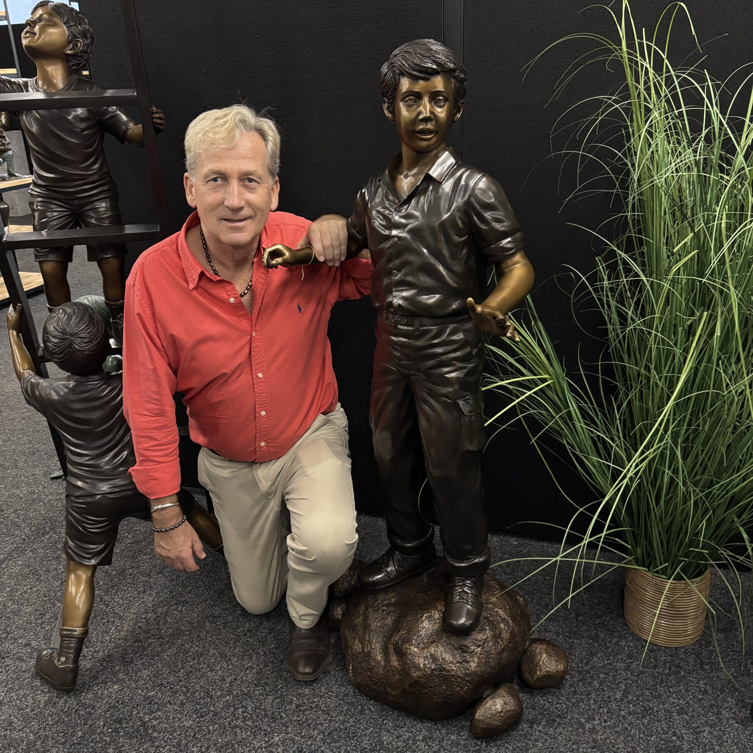 Bronze Young Boy Explorer Garden Sculpture FIBO 73 life-size bronze figure standing on textured rocks, photographed indoors beside ornamental grasses for scale and detail