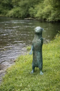 Curious Otter Bronze Garden Sculpture Fountain Water Feature WI 117 standing naturally beside a river in a waterside garden setting.